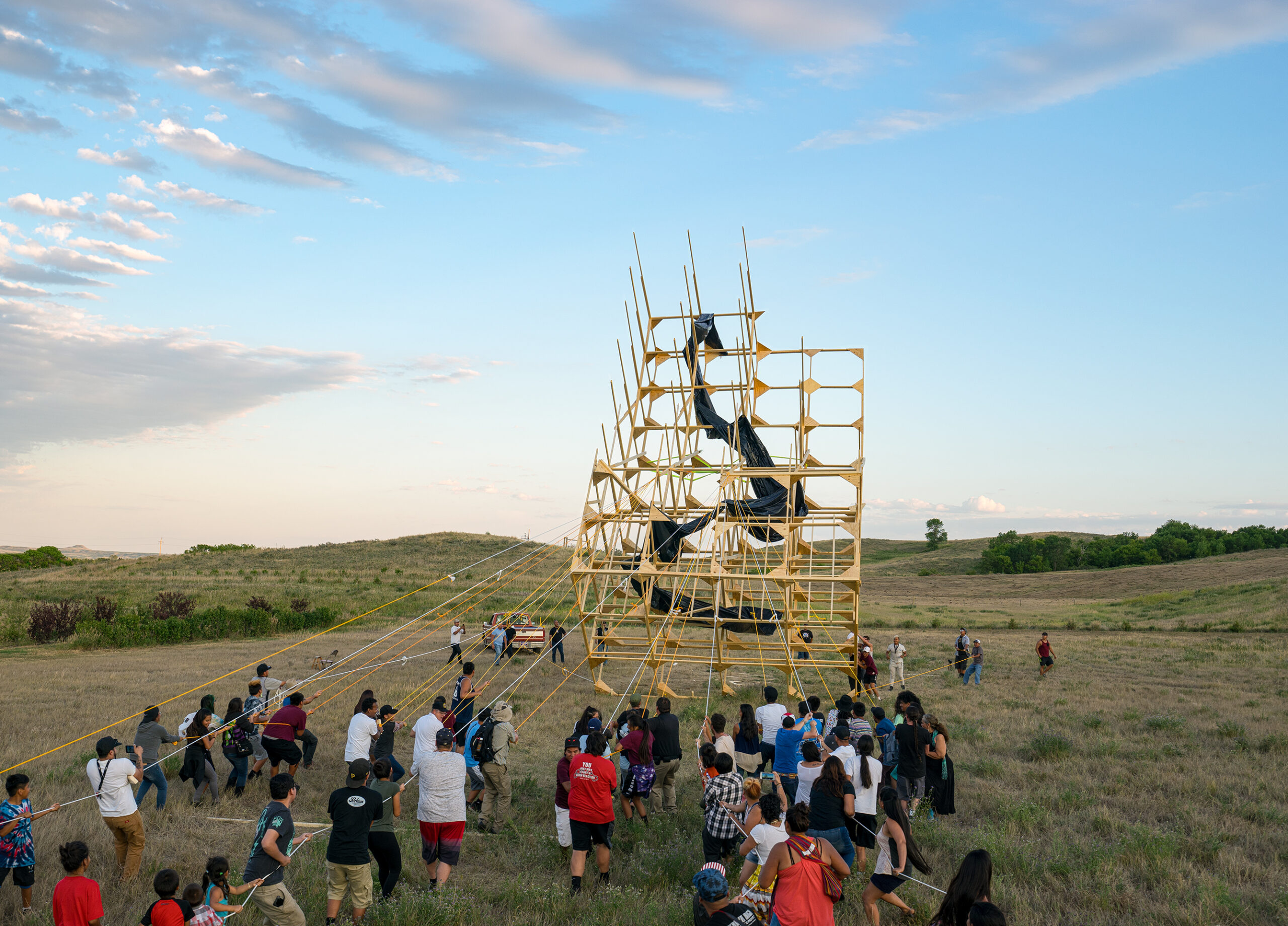 【写真】広い草原の奥側に黄色い大きな構造体があり、その構造体から伸びた多くのロープを手前側にいる50人近くの人がそれぞれ握っている
